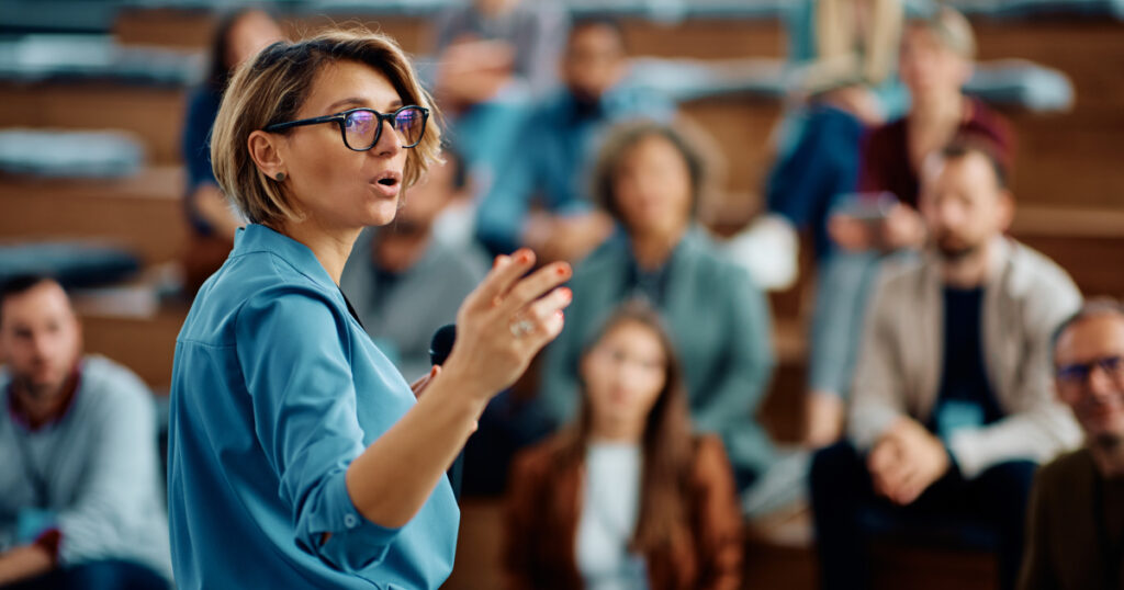 Person holding a microphone giving a talk to a room of people in the background