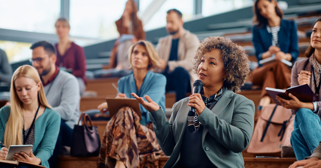 Person holding a microphone talking with a people sitting in the background in a lecture theatre